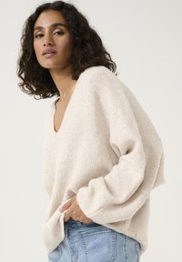 Pull beige confortable avec un décolleté en V, coupe ample et accents scintillants subtils, associé à un jean en denim bleu clair.