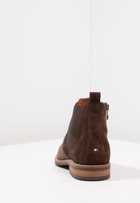 Stivale alla caviglia in suede marrone con pannelli laterali elasticizzati, chiusura con zip e tacco in legno a strati. Presenta una piccola etichetta con logo sul retro.