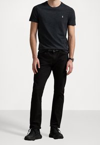 Man med mörkgrå kortärmad skjorta, svarta jeans, svart bälte, svarta sneakers och armbandsur, står mot en enfärgad vit bakgrund.