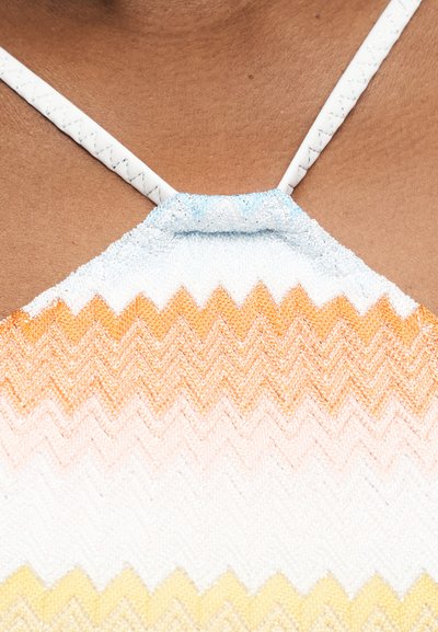 Κοντινή λήψη ενός υφασμάτινου τοπ με λευκές τιράντες και ζιγκ-ζαγκ μοτίβα σε πορτοκαλί, λευκό και κίτρινο πάνω σε καστανό τόνο δέρματος.