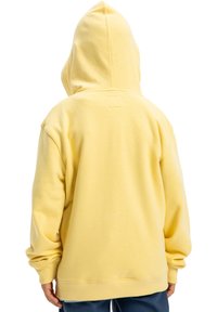 Osoba w luźnej, żółtej bluzie z kapturem, widziana z tyłu, w ciemnoniebieskich spodniach.