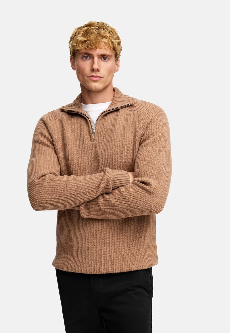 Halvfodrad stickad tröja i brunt, med ribbat mönster, långa ärmar och rak fåll. Bar över en vit T-shirt och matchad med svarta byxor.