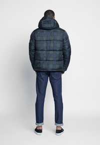 En person står vänd bort, iklädd en huva, puffy rutjacka, mörka jeans och svarta skor, mot en enkel vit bakgrund.