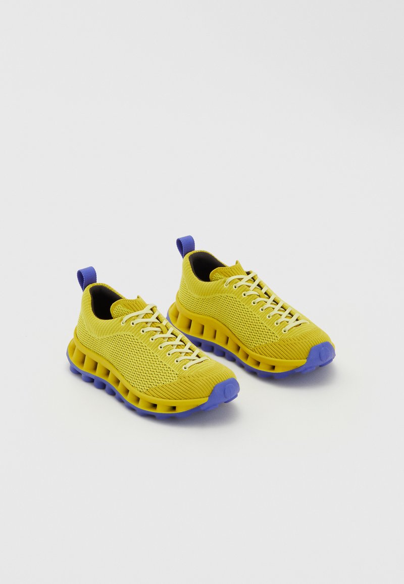 Ténis de malha amarelos com sola e detalhes roxos, apresentando uma sola almofadada, fecho com atacadores e uma peça na parte de trás para calçar facilmente.