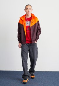 Chaqueta con capucha de color marrón oscuro y naranja, camiseta gráfica roja, jeans grises y zapatillas negras con naranja; de pie contra un fondo sencillo.