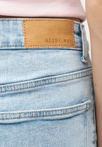 Φωτεινά γαλάζια τζιν παντελόνια από denim με καφέ δερμάτινο ένθετο με την ετικέτα «NOISY MAY», φορεμένα από ένα άτομο με ροζ κορυφή, εμφανιζόμενα από την πλευρά της μέσης.