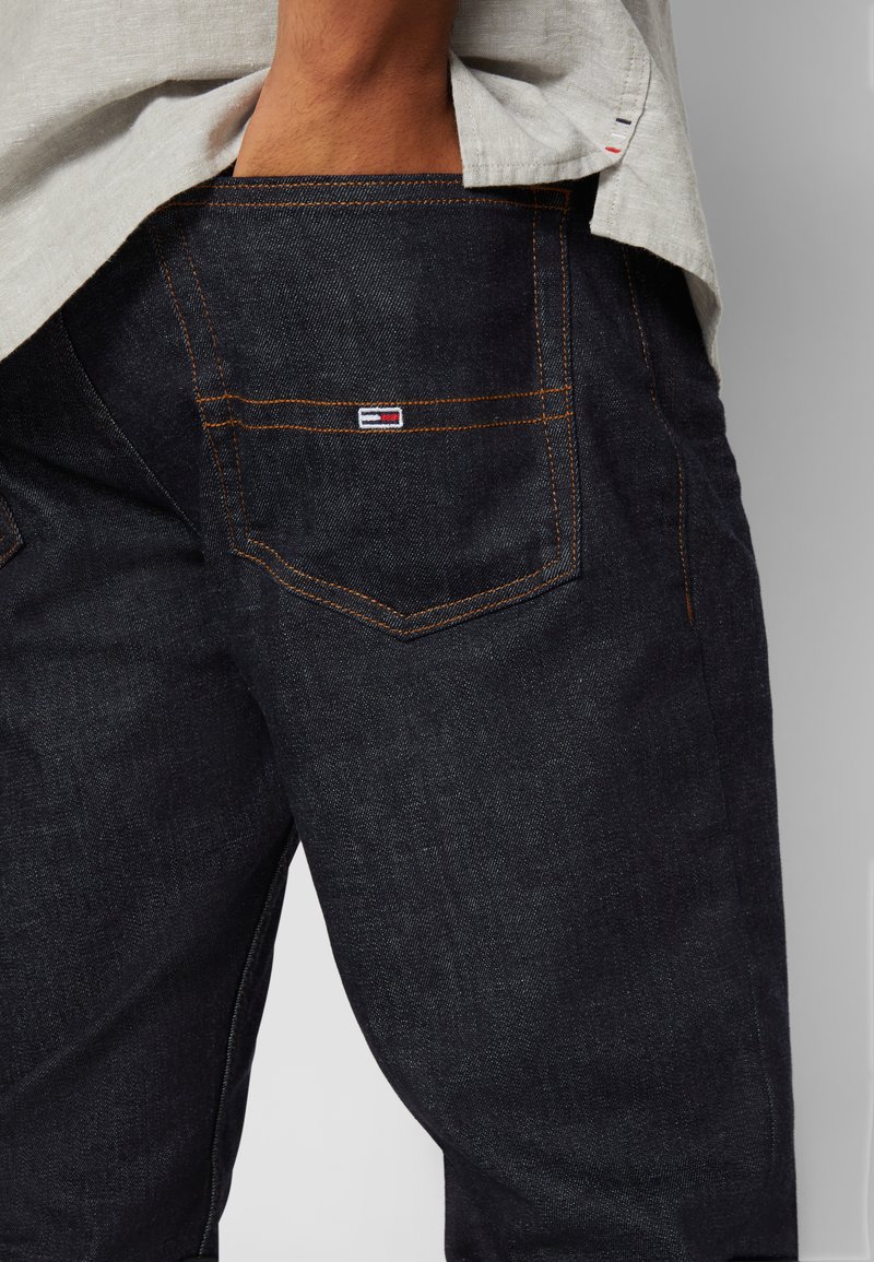 Donkerblauwe denim jeans met oranje stiksels, met een achterzak met een klein logo-opplekje. De stof heeft een gladde textuur.