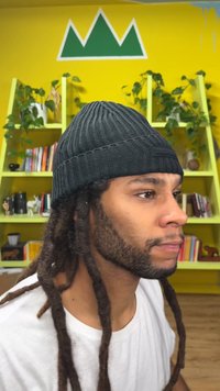 Schwarze Strickmütze mit geripptem Muster, getragen von einer Person mit Dreadlocks und weißem Hemd, vor einem leuchtend gelben Hintergrund mit grünen Akzenten.
