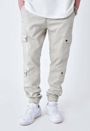 Personne portant un pantalon cargo beige clair avec des poignets élastiques et des baskets blanches, se tenant avec les mains dans les poches sur fond blanc.