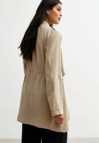 Blazer beige a maniche lunghe con vita cintrata, texture liscia, tasche laterali e polsini con bottoni. Retro semplice con dettagli di cucitura minimal.