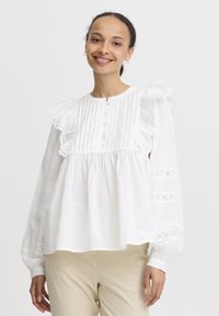 Bijela pamučna bluza s volanima na rukavima, vertikalnim nabranim detaljima i zatvaranjem na gumbe. Ima detalje od čipke i opušteni kroj. Dugi rukavi.