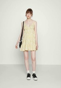 Vestido floral amarelo com decote em V, alças finas e saia evasê. Usado com ténis pretos e meias para o tornozelo, acompanhado de uma mala preta.