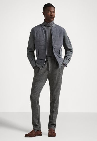 Mężczyzna ubrany w szarą pikowaną kurtkę bomberkę, szary golf, szare spodnie i brązowe zamszowe mokasyny, stoi z rękami w kieszeniach.