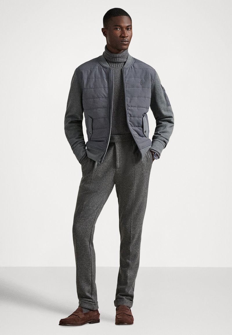 Homme portant une veste bomber matelassée grise, un pull à col roulé gris, un pantalon gris et des mocassins en daim marron, debout les mains dans les poches.