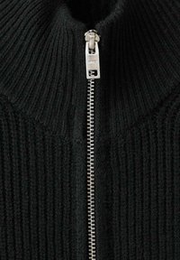 Detailný záber na strieborný zips na čiernej ryhovanej pletenine, zobrazený čiastočne rozopnutý v strede.