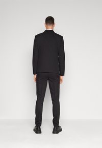 Black suit with tailored jacket and straight-leg trousers. Smooth fabric, minimal detailing. Worn with black shoes, viewed from the back.