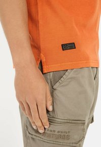 Oranje T-shirt met korte mouwen en zijsplitten, gemaakt van een textuurstof. Gecombineerd met kaki cargoshorts met voor- en zijzakken.