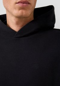 Ritratto ravvicinato di una persona che indossa un felpa nera con cappuccio, sulla quale è ricamato un piccolo logo "QS" sul petto.