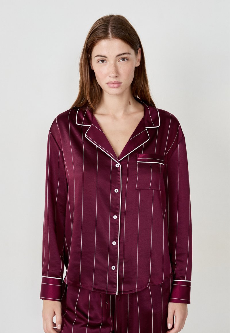 Burgundowa satynowa bluzka piżamowa z białymi paskami, długimi rękawami, kieszenią i zapięciem na guziki, z kontrastującymi białymi lamówkami przy kołnierzyku i mankietach.