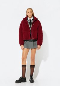 Burgunderfarbene Kordjacke mit versteckten Knöpfen, grauer Faltenrock, schwarze Stiefeletten und kniehohe schwarze Socken.