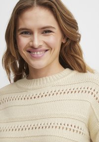 Maglione lavorato a maglia beige chiaro con collo rotondo a coste, caratterizzato da righe orizzontali e motivi decorativi a maglia aperta. Texture morbida.