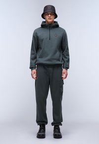 Mørkegrøn hoody med frontlynlås og lille logo, parret med matchende sweatpants med cargo lommer og elastiske mansjetter. Sorte sko og en bucket hat.