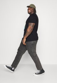Black t-shirt with graphic design, gray jeans, and black high-top sneakers. Olive green cap. Person walking, smiling, viewed from the side.
