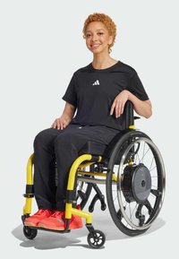 Maglietta e pantaloni sportivi neri indossati da una persona in una sedia a rotelle gialla. Scarpe da ginnastica rosse vivaci. Texture liscia e design moderno.