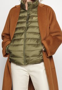 Persona vestida con un abrigo de lana marrón sobre una chaqueta acolchada verde oliva con el logo "BOSS", combinado con pantalones blanco roto, con las manos en los bolsillos de la chaqueta.