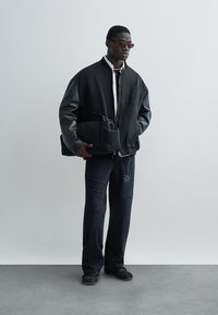 Μαύρο μπουφάν bomber με δερμάτινα μανίκια, λευκό πουκάμισο, σκούρο τζιν με πατς, μαύρα αθλητικά παπούτσια και μία δομημένη μαύρη τσάντα.