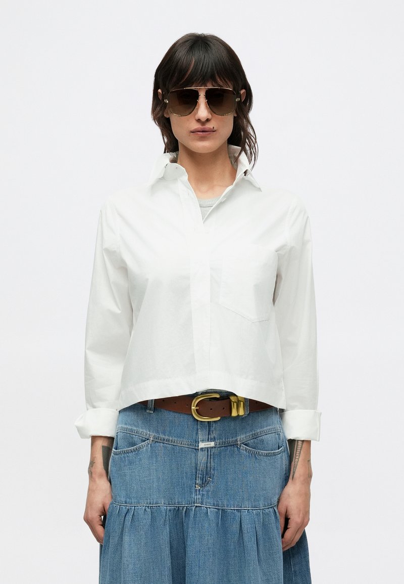 Biela skrátená košeľa s dlhými rukávmi a náprsným vreckom, v kombinácii s rifľovou sukňou s volánovým lemom a hnedým opaskom so zlatou prackou.