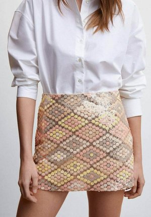 Kobieta w białej koszuli zapinanej na guziki z podwiniętymi rękawami oraz wysokiej talii mini spódnicy z cekinami i pastelowymi geometrycznymi wzorami.