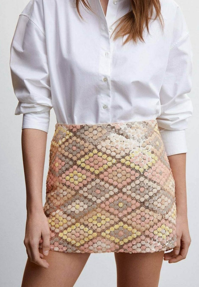 Femme portant une chemise blanche boutonnée avec les manches retroussées et une mini-jupe à sequins taille haute avec des motifs géométriques pastel.