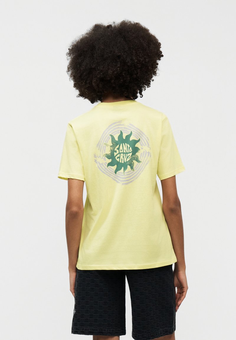 Άτομο με σγουρά μαλλιά φοράει κίτρινη μπλούζα με πράσινο ηλιακό γραφικό "Santa Cruz" στην πλάτη και μαύρο σορτς με υφή, στέκεται μπροστά σε λευκό φόντο.