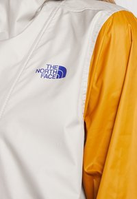 Vit och gul jacka med en blank yta. Funktioner inkluderar en blå The North Face-logotyp fram och ärmar i kontrasterande färg.