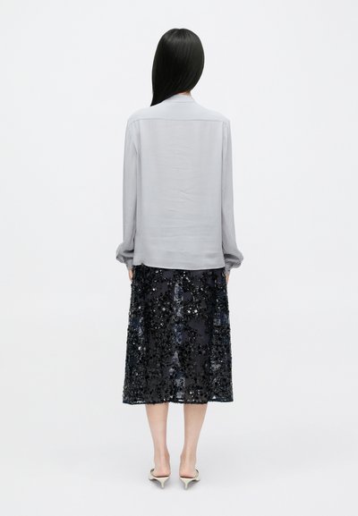 Kobieta z długimi czarnymi włosami stoi tyłem, ubrana w jasnoszarą bluzkę z długim rękawem, czarną błyszczącą spódnicę midi oraz białe buty na obcasie.