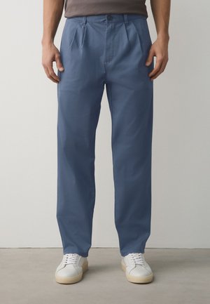 Mann trägt blaue gerade geschnittene Hose und weiße Sneaker und steht vor einem schlichten hellen Hintergrund.