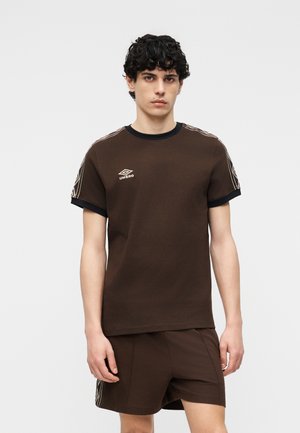 Νεαρός άνδρας όρθιος, φορεμένος με καφέ κοντομάνικη μπλούζα Umbro και ταιριαστά σορτς με μαύρα περιθώρια και λευκές ρίγες στα μανίκια και στις πλευρές.