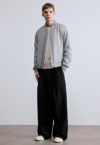 Világosszürke bomberdzseki cipzáros előlappal, bordázott mandzsettákkal és texturált felülettel, bézs pulóver és fekete, bőszárú nadrág fölött viselve. Fehér cipők.