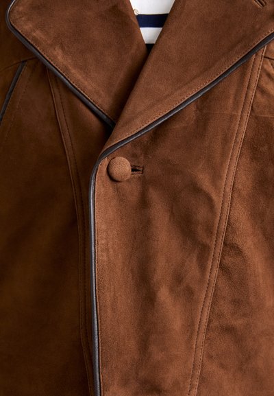 Cappotto in suede marrone con ampio colletto, piping nero a contrasto, unico bottone testurizzato e dettagli di cucitura visibili. Tessuto morbido e liscio.