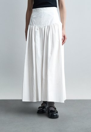 Žena nosi dlhú bielu sukňu so širokým pásom, tmavé ponožky, čierne topánky so sponou a čierne tielko bez rukávov.