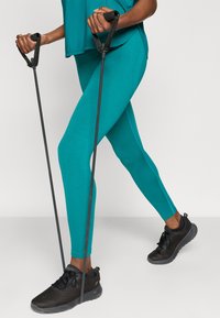 Τουρκουάζ αθλητική εμφάνιση με άνετο τοπ και εφαρμοστά κολάν. Μαύρες λωρίδες γυμναστικής κρατιούνται στα χέρια, συνδυασμένες με μαύρα αθλητικά παπούτσια.