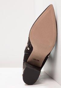 Musta mokkanahkainen korkokenkä, jossa on terävä kärki, koristeelliset kultaiset niitit ja sileä vaaleabeige pohja, jossa on uurteinen pinta.