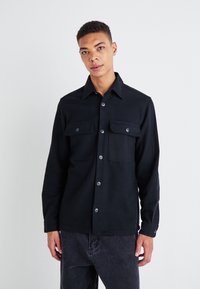 Chemise noire à boutons avec un col pointu, deux poches poitrine et des manches longues. Fabriquée à partir d'un matériau texturé avec une finition lisse.