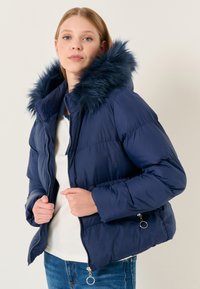 Giubbotto puffer blu navy con cappuccio in pelliccia sintetica, texture trapuntata, chiusura frontale con zip e tasche laterali con zip, indossato sopra una maglietta bianca.