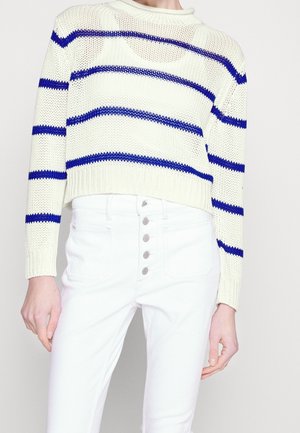 Person iført en cremeknitsweater med horisontale blå striber og hvide jeans med høj talje med fem synlige knapper foran.