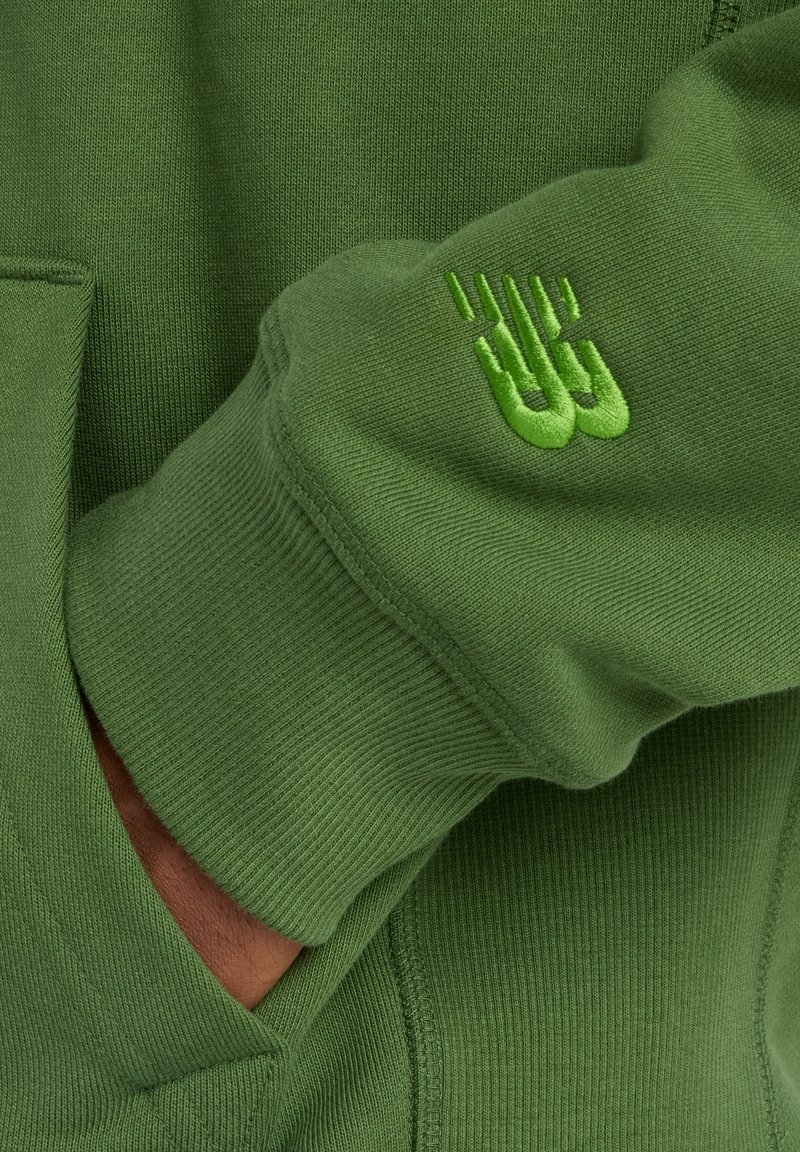 Sweat-shirt vert avec poignets côtelés et finition texturée. Logo brodé notable en vert clair sur la manche.