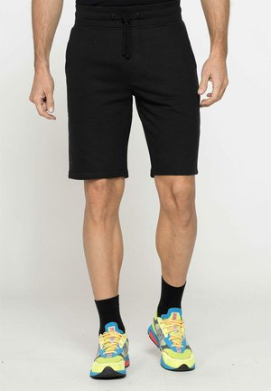 Uomo che indossa pantaloncini neri con coulisse, calzini neri e scarpe da ginnastica colorate, in piedi davanti a uno sfondo uniforme.