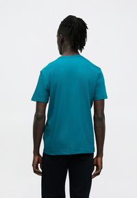 Modré bavlnené tričko s voľným strihom, krátkymi rukávmi a okrúhlym výstrihom, pohľad zozadu. Jednoduchý dizajn, bez vzorov alebo ozdôb.