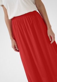 Jupe maxi rouge avec une taille élastique, présentant un tissu fluide et texturé qui drape doucement. Portée avec un t-shirt blanc.